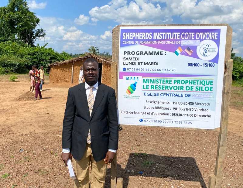A native of Ivory Coast, a student, is standing next to a buildboard of Shepherds Institute
